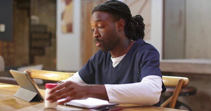 African American man in scrubs at table using tablet lifting red-lid bottle checking label noting