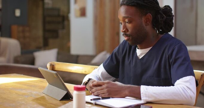 African American man in scrubs joining tablet call for telehealth, writing notes at dining table