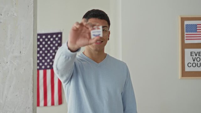 Young hispanic man holds 'voted' sticker between fingers, smiling, in a polling building with american flag visible; civic pride.