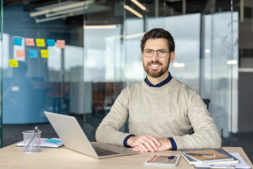 Fototapeta premium Smiling bearded professional wearing glasses sits at a modern desk with laptop and office supplies, looking confident and relaxed while working remotely in a contemporary workspace