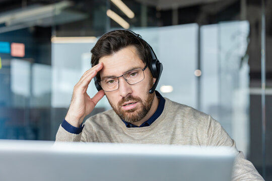 Overworked male call center agent experiencing stress and a headache during a difficult customer support call, burnout affecting his concentration in a modern office environment
