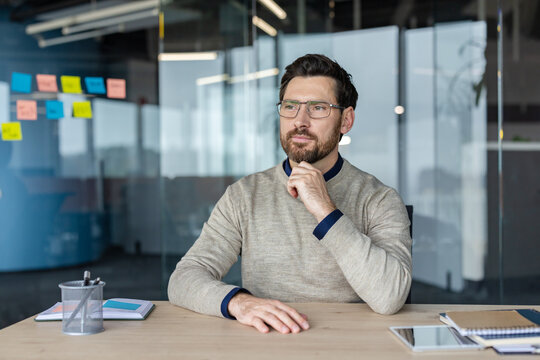 Confident bearded businessman wearing eyeglasses sitting at desk, deep in thought and planning future strategy while working in a contemporary office environment