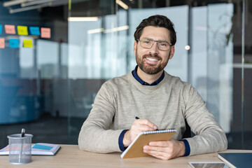 Man with glasses and beard smiling at the camera while sitting at a desk, writing in a notepad,...
