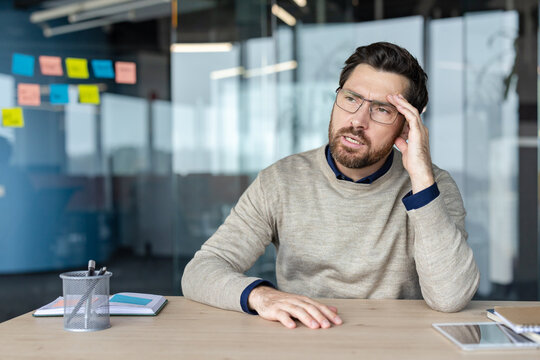 Worried professional man experiencing stress and a painful headache while sitting at an office desk, feeling overwhelmed by problems and work pressure