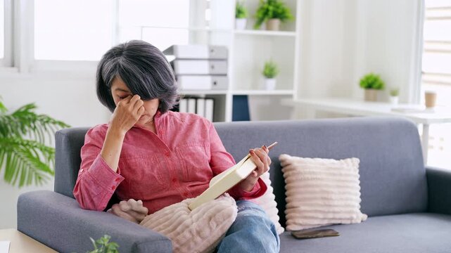Senior Asian woman suffering eye strain while reading book at home  Elderly lifestyle concept, vision problem, stress and health issue.