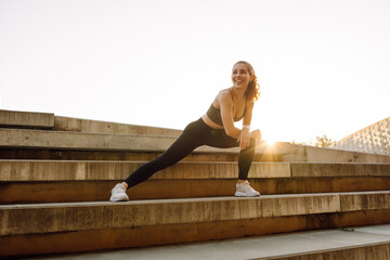 Naklejka premium An athletic woman exercises on the steps on a sunny day. A young woman in athletic clothing stretches while exercising outdoors at sunset. Concept of activity, sports, and weekends.