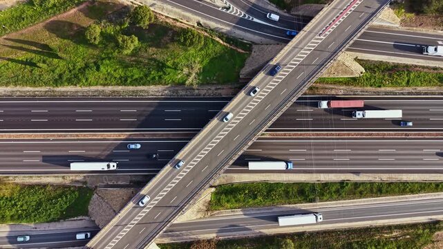 Cars move along highways while trucks travel over an intersection in a rural setting. Green fields and trees surround the roads under a clear sky.