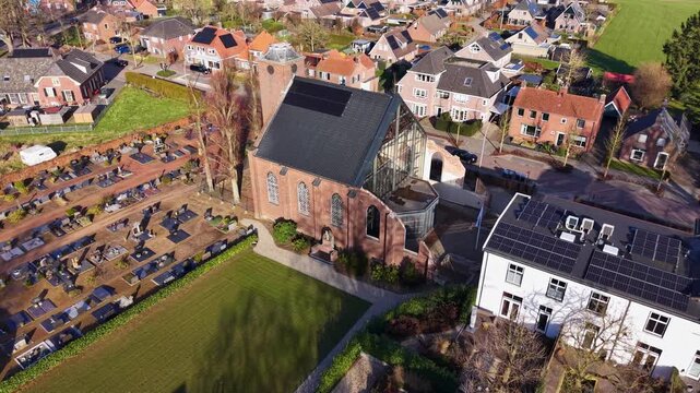 Aerial view of a European village featuring a historic brick church with a modern glass nave, an adjacent cemetery, and residential houses with solar panels bordering green fields in sunlight.
