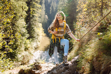A woman stands on a narrow path by a river. She holds a rope for balance and looks around. Green trees surround her in the forest. This scene is vibrant and lively. Hiking concept. © maxbelchenko