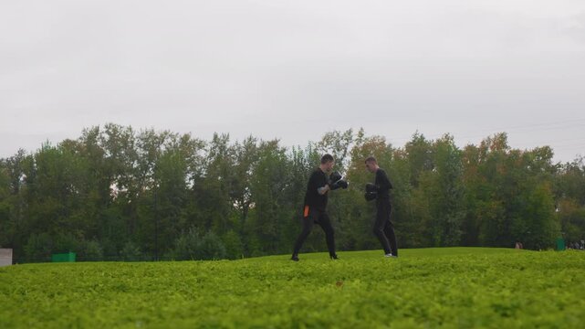 two men practicing kicks in field during energetic sparring drill, fast footwork, partner exchanges, coach presence, damp grass and grey sky, focused faces, high intensity training