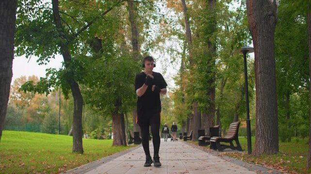 Caucasian man jogging down treelined path, wearing black training clothes, benches and fallen leaves lining sidewalk, steady stride, autumn light, calm focused expression, urban park scenery,