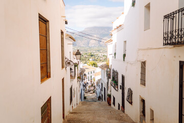 Fototapeta premium A narrow street leads through a small village with white buildings on both sides. The area has plants on balconies and mountains in the background under a clear sky.