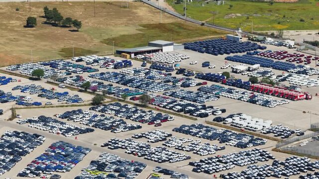 Impressive aerial perspective flying over a vast industrial parking lot filled with new cars, gradually revealing a scenic mountain landscape with sustainable wind turbines in the distance