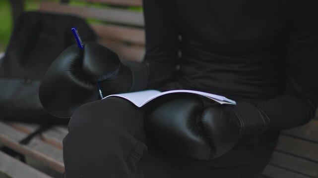 Caucasian athlete with gloves writing on paper while seated on park bench, closeup of leather mitts on lap and pen tracing strategy lines, calm daylight atmosphere, ritual preparation before training