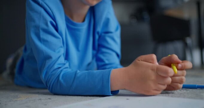 Young child in blue shirt concentrating while drawing with a yellow pencil on paper at home, close-up of hand and thoughtful face