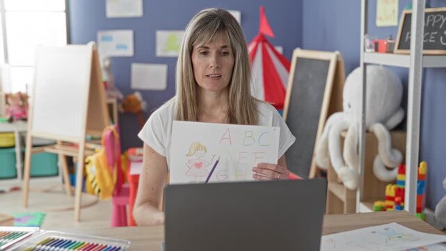 Woman holds alphabet drawing up to laptop while seated at table with crayons and toys in a playroom building; focused learning.