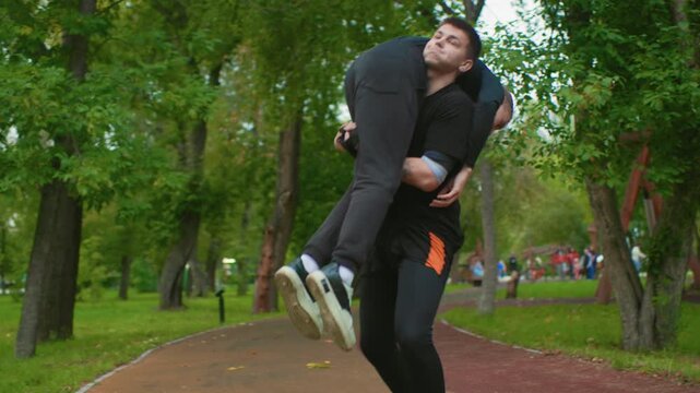 men sparring on park path during takedown training, trainer and trainee exchange punches and grappling moves, man thrown to ground on wet pavement, leafy trees and walking trail in background, focused
