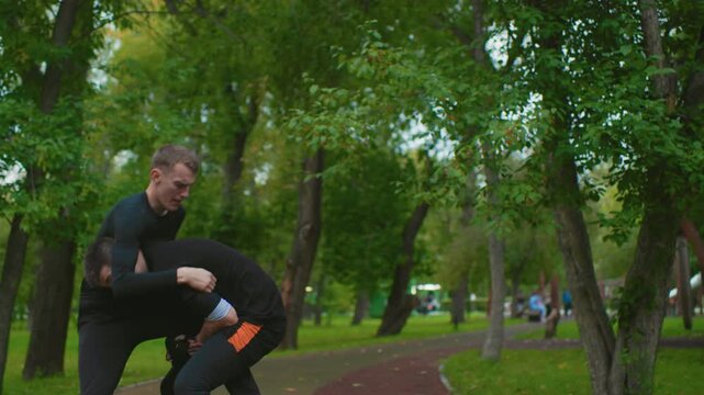white male fighters reconciling in leafy park after intense training, handshake and calm discussion between partners, coach nodding as duo walk off pathway, camaraderie and sportsmanship replacing