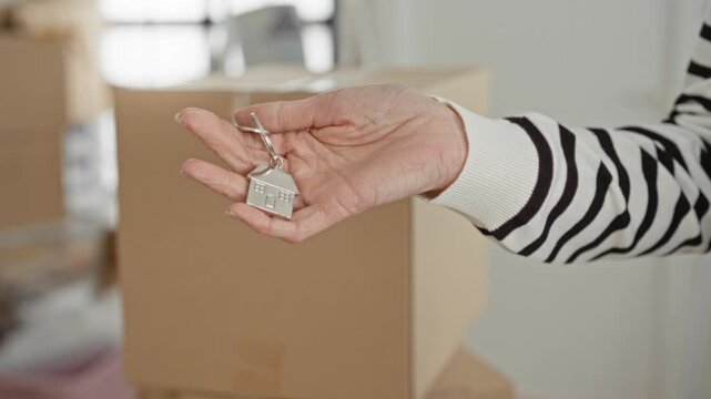 Woman holding a house key on a small house keychain, hand extended over stacked cardboard moving boxes inside a building; moving transition hope.