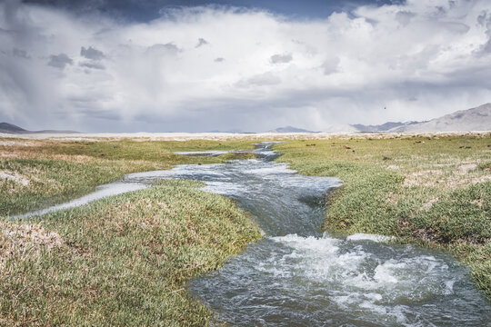 High in the Pamir Mountains of Tajikistan, a river flows along a raft with grassy banks against a backdrop of snow-capped mountain peaks. A panoramic view of the Tien Shan highlands