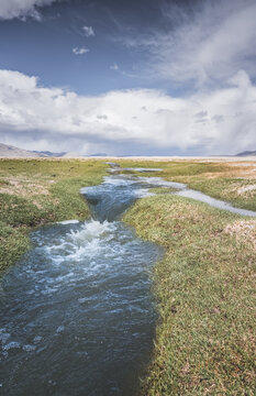 High in the Pamir Mountains of Tajikistan, a river flows along a raft with grassy banks against a backdrop of snow-capped mountain peaks. A panoramic view of the Tien Shan highlands