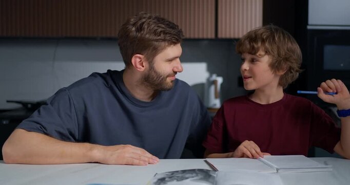 Father Offers Nurturing Assistance And Encouragement To Son During Study Session At Kitchen Table