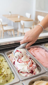 Close up of a female hand using a metal scoop to serve fresh berry swirl gelato in a professional ice cream shop. Pistachio and strawberry flavors are visible in metal container