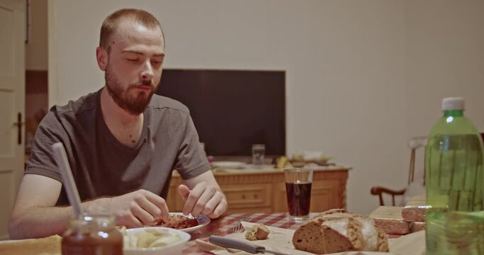 A young bearded man enjoys a simple meal at a wooden dining table in his home. He is focused on his food, tearing off pieces of bread, with a glass of dark drink beside him.