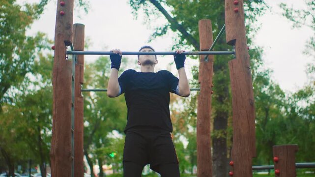 Pullups performed in park, Outdoor workout with intense arm effort, Man strains during pullups in leafy park setting, Solitary man engages in strenuous pullup exercise among trees in park