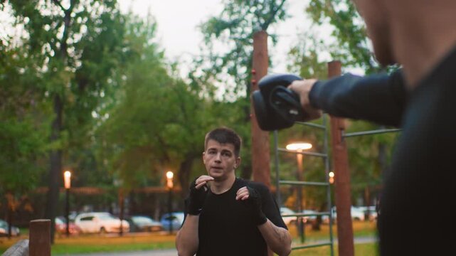 young man training with focus mitts in park at dusk showing defensive reaction and concentration coach throws measured punches to mitt, trainee raises guard, practices stance and counter technique