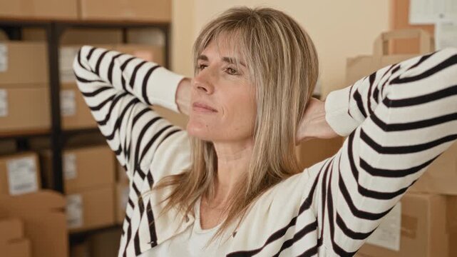 Woman stretching with hands behind head surrounded by cardboard packages in a building; small business relief.
