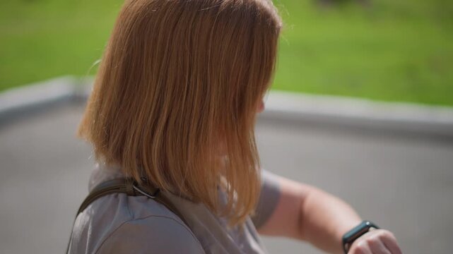 Back view of worried lady tapping smartwatch to check time under bright sunlight, golden hair glowing, carrying back strap, greenery blurred in background