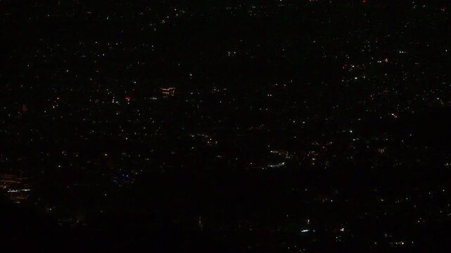 A close-up night view of the valley reveals a dense scatter of twinkling city lights, with faint outlines of buildings and curving roads glowing softly against the dark Mussoorie hills.