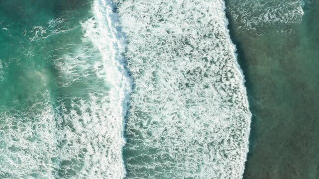top-down aerial footage of powerful ocean waves rolling across turquoise water and crashing toward the shoreline. The mesmerizing white sea foam patterns create a calming and cinematic coastal texture