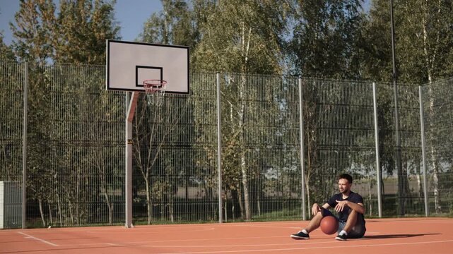 Lonely bored man sitting on sunny basketball court and bouncing balls with hands. Lonely amateur athlete waiting for friends to arrive, wide shot. Concept of loneliness, introvert, depression.