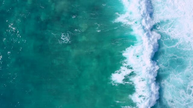 Top-down aerial footage of an ocean wave rolling and breaking into bright white foam over blue-turquoise water. The natural patterns and dynamic motion create a clean, modern adventurer 