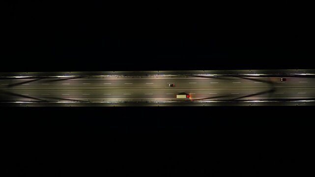Stunning aerial top-down perspective following vehicles traveling along a brightly lit highway bridge at night, revealing the road's linear structure against the surrounding darkness