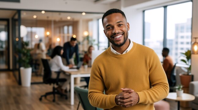 Smiling Black man in a yellow sweater standing in a modern office. Confident creative professional in a casual corporate workplace. Teamwork concept