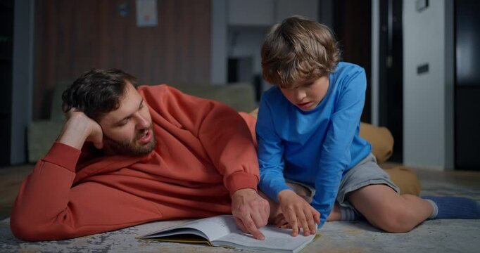 Father and young son lying on the living room floor reading a book together, sharing a quiet learning moment during homework at home
