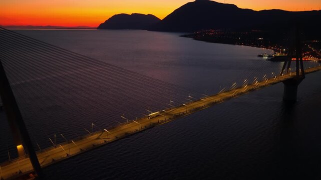 Stunning aerial view of a illuminated bridge at dusk, with white truck crossing the gulf of corinth against a backdrop of mountains and a vibrant orange sunset.