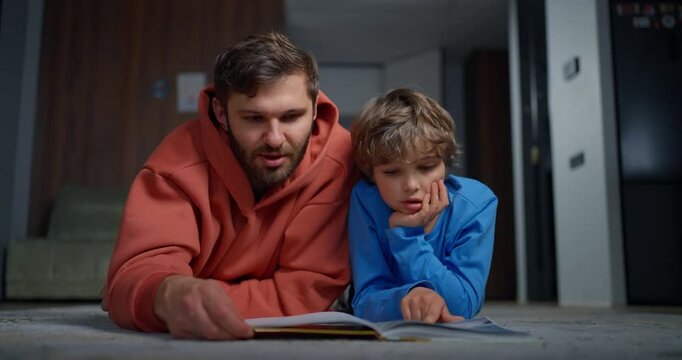 Father and young son lying on living room floor reading a picture book together, sharing a quiet bedtime learning moment at home