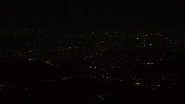 A dimly lit night view from Mussoorie shows a sprawling valley sprinkled with faint, scattered city lights and barely visible winding roads fading into the dark hills.
