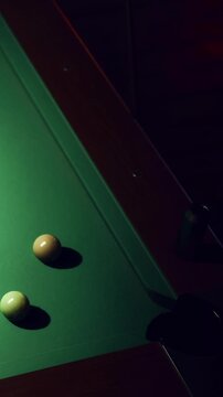 A person is aiming a cue stick at a billiard ball on a green pool table in a dark room. The focus is on the cue ball and the anticipation of a shot.