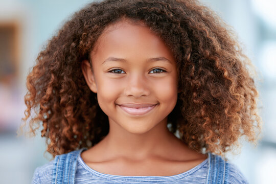Beautiful young girl with curly brown hair smiling warmly looking directly at camera. Cute happy female child portrait showing positive emotion against soft blurry blue background with empty space