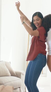 Vertical video: Smiling, dancing two women pressing back-to-back, swaying hips at home with sofa