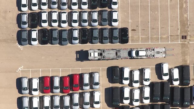 Overhead drone view as it ascends, revealing a car carrier driver unloading a vehicle and parking it into an empty spot among neat rows in a large port storage terminal.