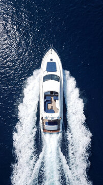 High angle aerial view of a luxury white motor yacht cruising through deep blue sea water. The boat leaves a white trail of foam and wake while sailing on a sunny day in the ocean.