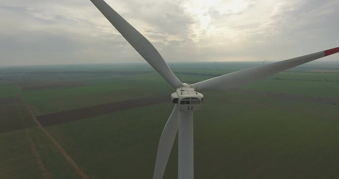 Drone orbits a large wind turbine at blade level above vast green agricultural fields under overcast sky. Nacelle marked 3.2, red-tipped safety blade, distant turbines visible on the horizon