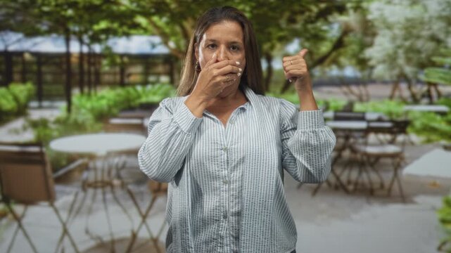 Woman covers mouth with hand and shows thumb up gesture at a street cafe patio among tables and chairs; surprise uncertainty.