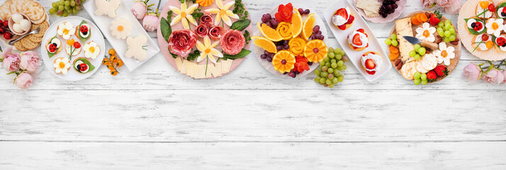 Naklejka premium Summer garden party food top border. Assortment of fun flower theme charcuterie items, fruit, crostini, finger sandwiches and dessert. Overhead view on a white wood banner background.